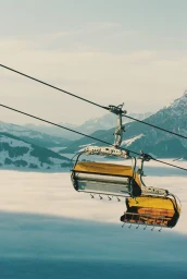 Yellow chairlifts suspended on steel cables above a sea of low clouds and snow-covered alpine mountains.
