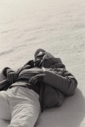 Model Lois Schindeler lying back on snow wearing a winter puffer jacket, knit sweater, ski trousers, and dark sunglasses.