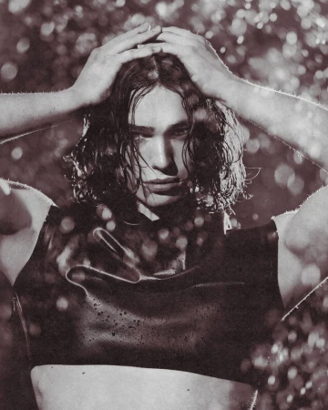 Portrait of a muscular adult male with wet dark hair, posing in a sleeveless shirt against a blurred, bokeh-filled backdrop.