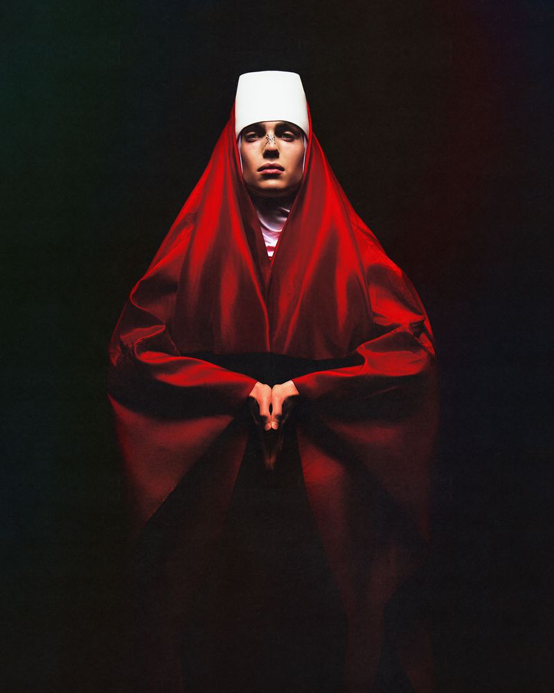 Person in red robe and white headwear stands in front of a dark background, hands clasped. Studio lighting highlights the form.