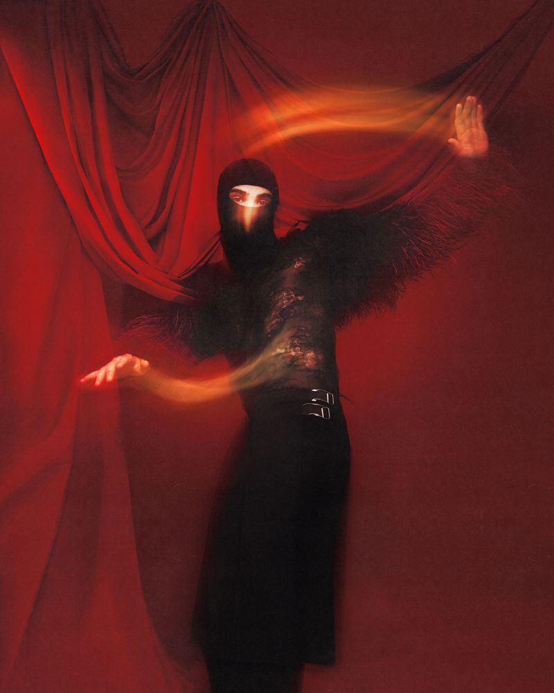 Person in a black outfit with fringed sleeves poses against a deep red backdrop with flowing light effects, creating a dramatic image.