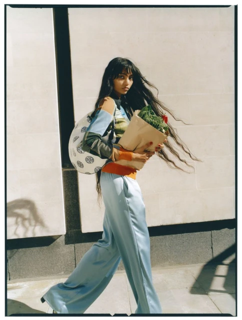 A stylish woman in flowing blue pants and a colorful top walks along a city street carrying a tote bag and a brown paper bag with greens.