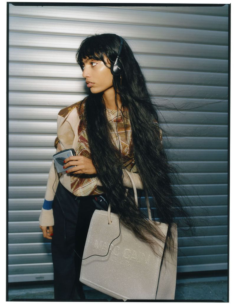 A woman with long dark hair, wearing a patterned top and a tote bag, listens to music on a portable music player against a metal wall.