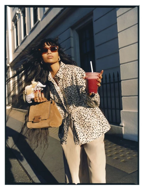 Stylish woman with long dark hair, wearing sunglasses and an animal print shirt, holds a smoothie and sandwich on a city street.