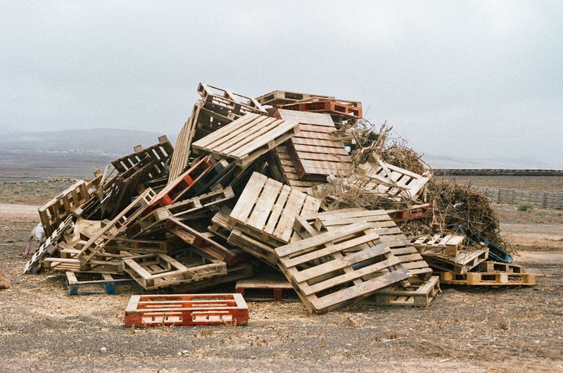 Large pile of wooden shipping pallets outdoors with a rural landscape and cloudy sky in the background, creating a textured scene.