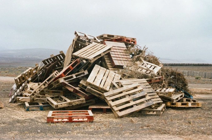 Large pile of wooden shipping pallets outdoors with a rural landscape and cloudy sky in the background, creating a textured scene.