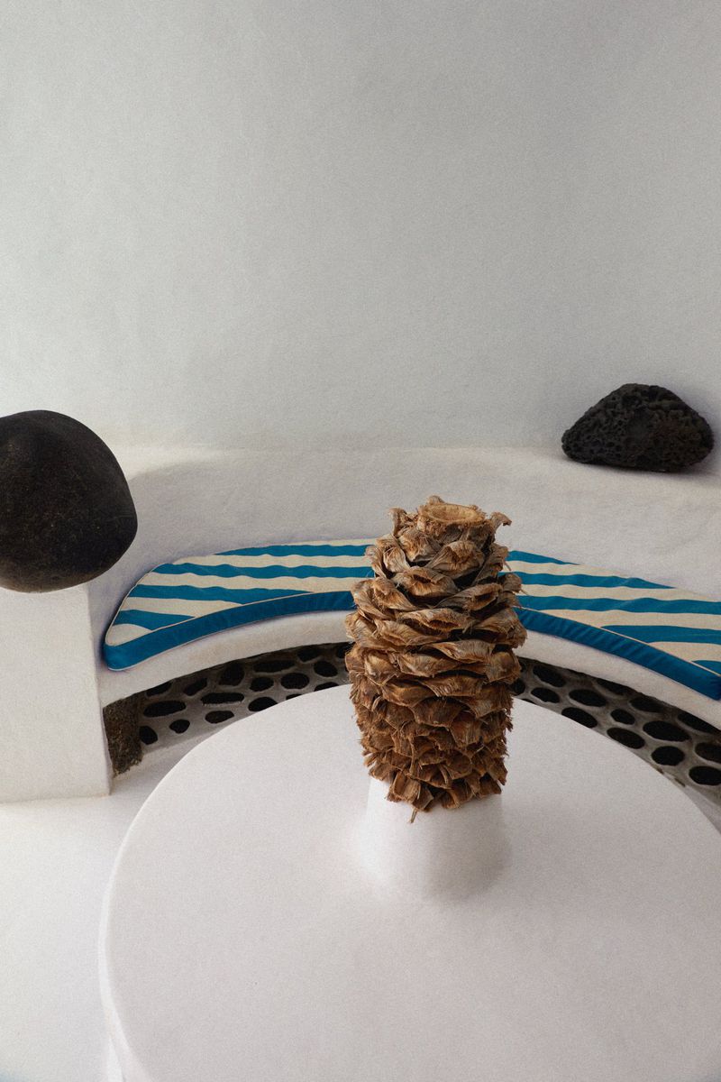 Still life featuring a pinecone on a white round table with rounded stones and a bench with blue and white stripes in the background.