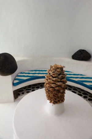 Still life featuring a pinecone on a white round table with rounded stones and a bench with blue and white stripes in the background.