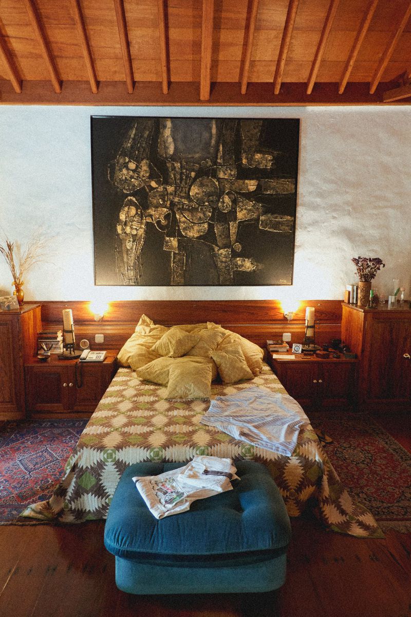 Bedroom interior featuring a queen-size bed with a patterned quilt, surrounded by wooden furniture, artwork, and a blue ottoman.