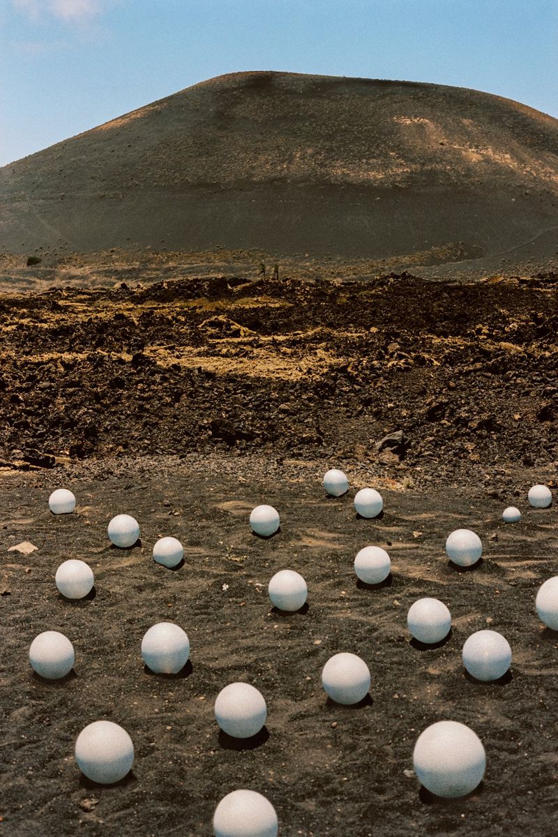 Landscape view of black rocky terrain scattered with white spheres, set against a backdrop of a dark, volcanic mountain under a clear sky.