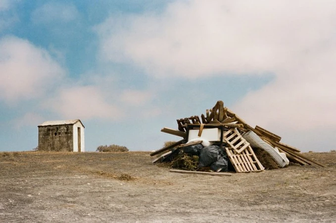 Wide shot of a debris pile in a barren landscape under a cloudy sky, with a small, weathered building visible in the distance.
