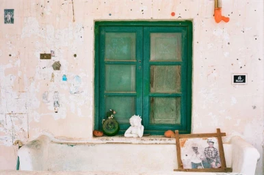 Weathered exterior wall with a green wooden shuttered window, a small cherub statue on the sill, and a framed photograph.