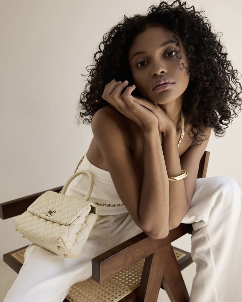 Portrait of an African American woman with curly hair, wearing a white outfit and sitting on a wooden chair, holding a purse.