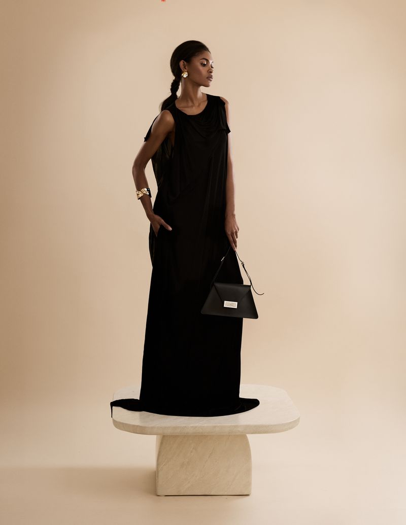 Elegant adult woman in a black dress and accessories, standing on a table with a purse, in a light studio setting.