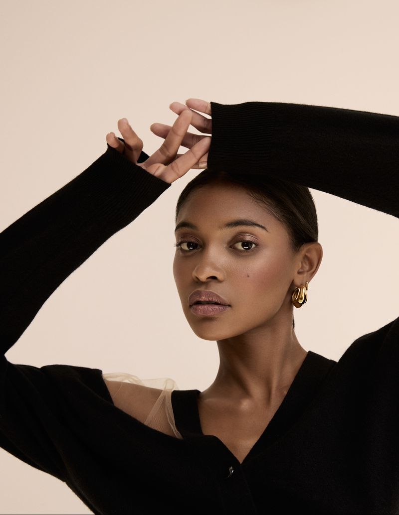 Portrait of a Black woman in a black V-neck sweater with sheer shoulder detail and gold hoop earrings, posing against a neutral background.