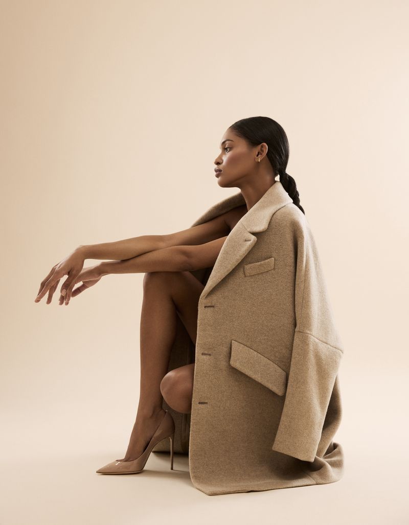 Elegant woman in a tan coat and matching high heels, seated against a neutral backdrop, exuding a minimalist fashion aesthetic.