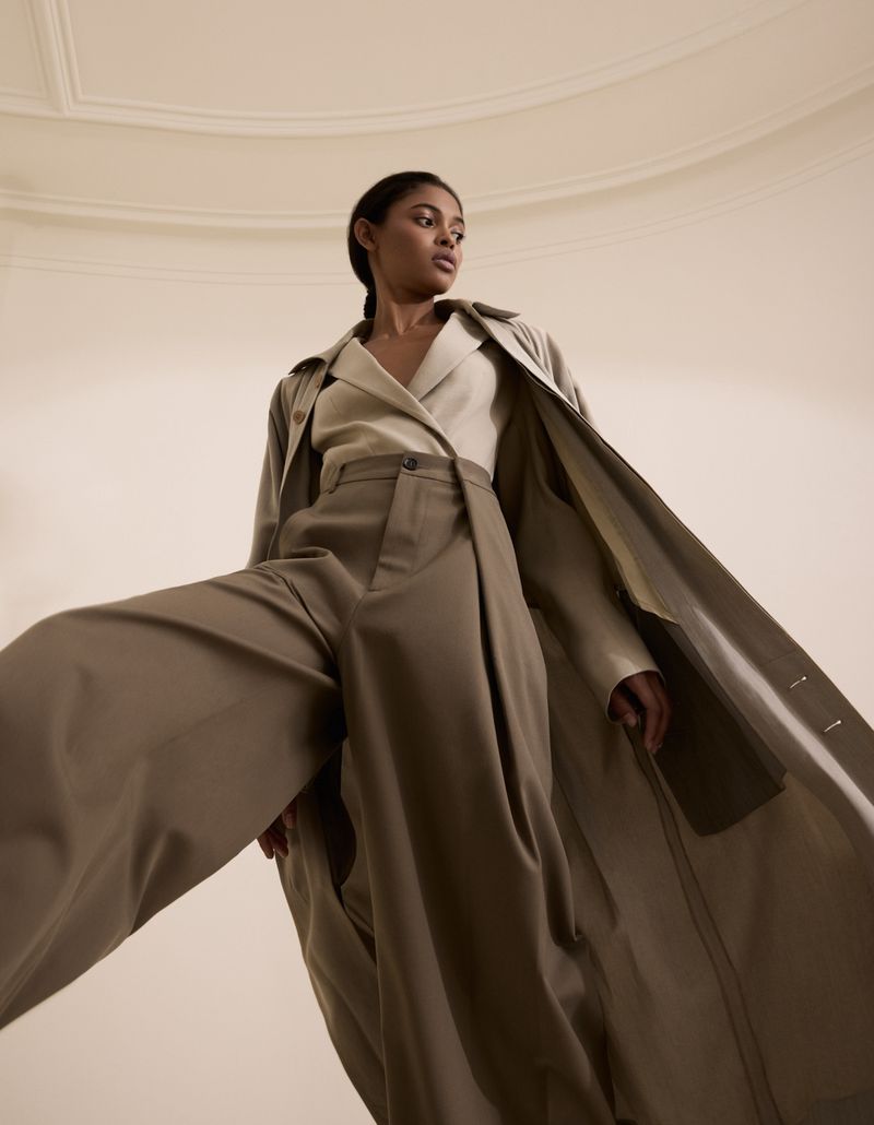 Fashionable woman posing in wide-leg trousers and a trench coat against a minimalist neutral backdrop, shot from a low angle.