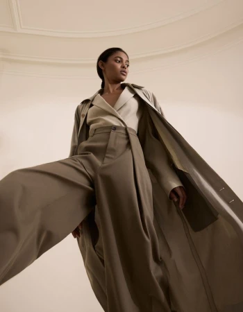 Fashionable woman posing in wide-leg trousers and a trench coat against a minimalist neutral backdrop, shot from a low angle.