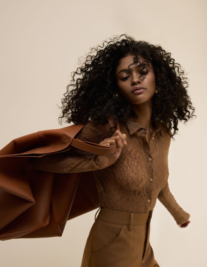 A woman with curly hair wearing a brown lace top and brown pants, carrying a large leather tote bag over her shoulder.
