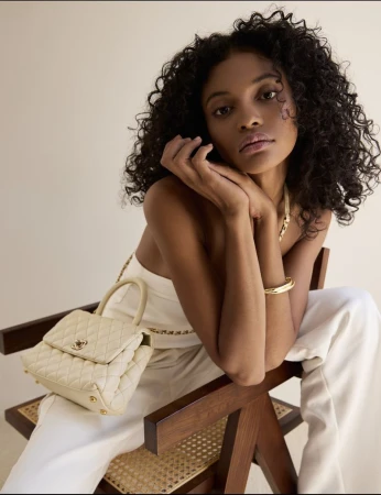 Model with curly hair seated on a wooden chair wearing an ivory outfit and holding a cream quilted top-handle handbag.