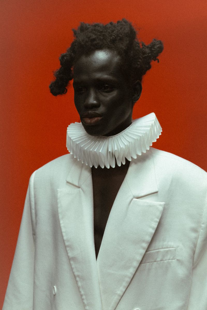 Portrait of a dark-skinned male in a white blazer and pleated neck ruff against a solid orange background in a photo studio.