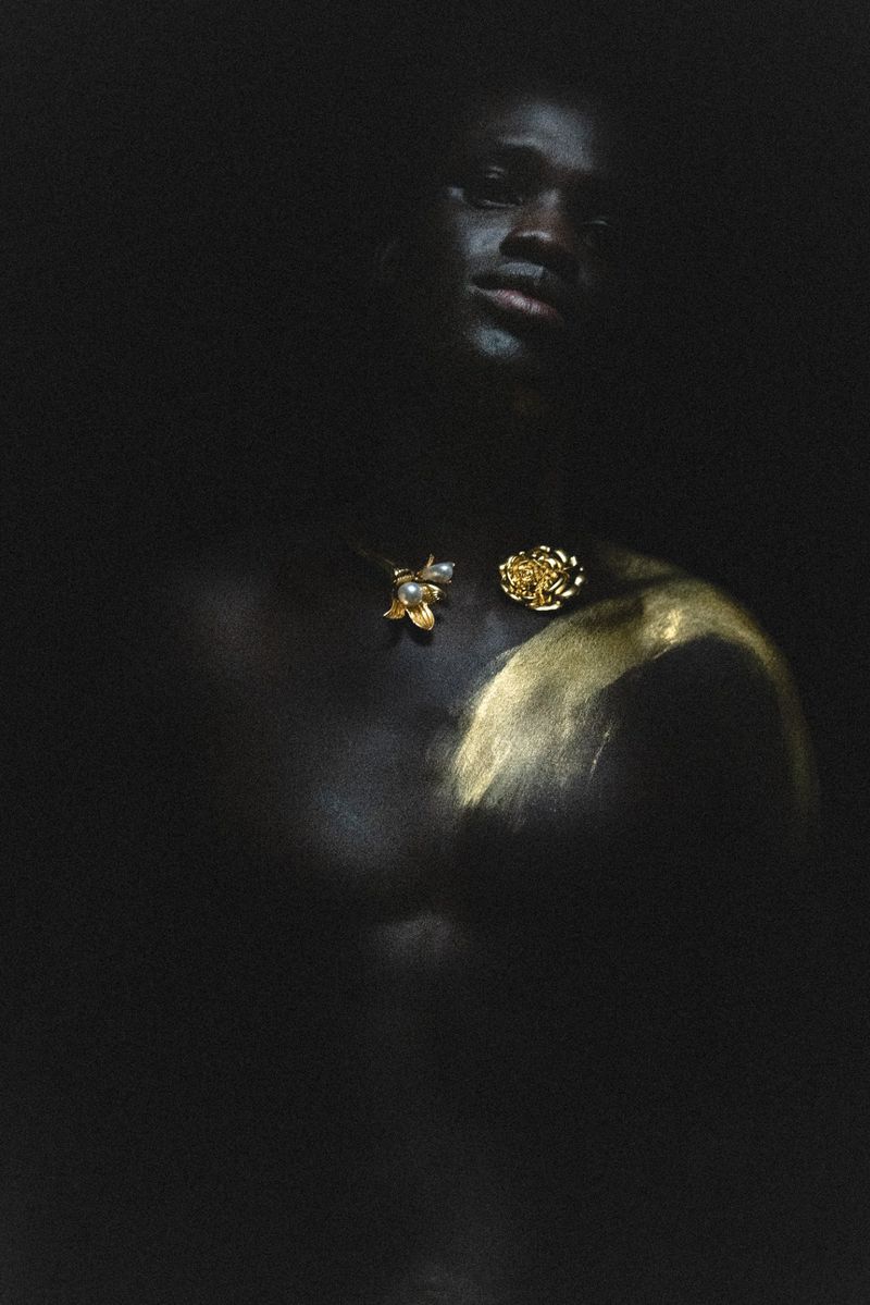 Portrait of a shirtless Black man with a confident expression, adorned with gold body paint and antique jewelry against a dark backdrop.