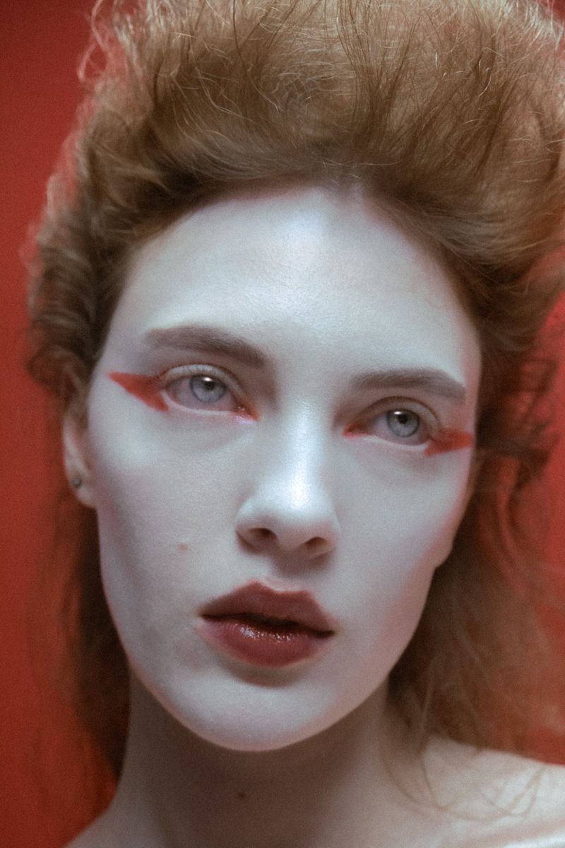 Close-up portrait of a woman with painted white skin, red eye makeup, and dark red lipstick against a solid red background.