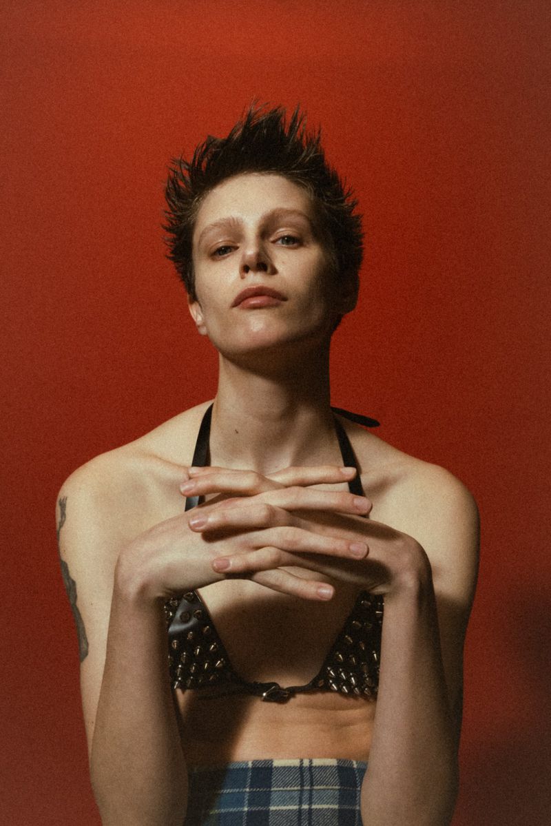 Close-up studio shot of a model wearing a studded bikini top and plaid skirt, posing with hands clasped, against a red backdrop.