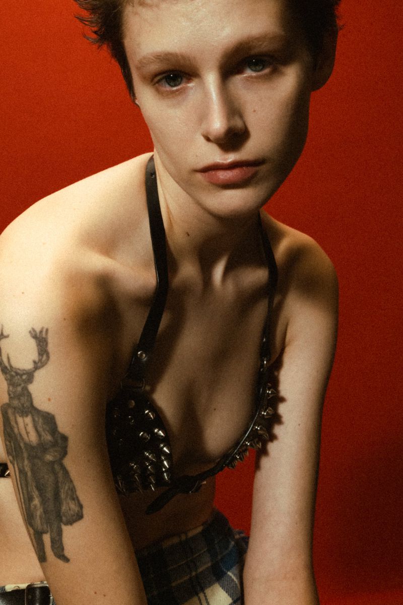 Close-up of a young adult woman with short hair and a tattoo on her arm wearing a studded bra and plaid bottoms against a red backdrop.