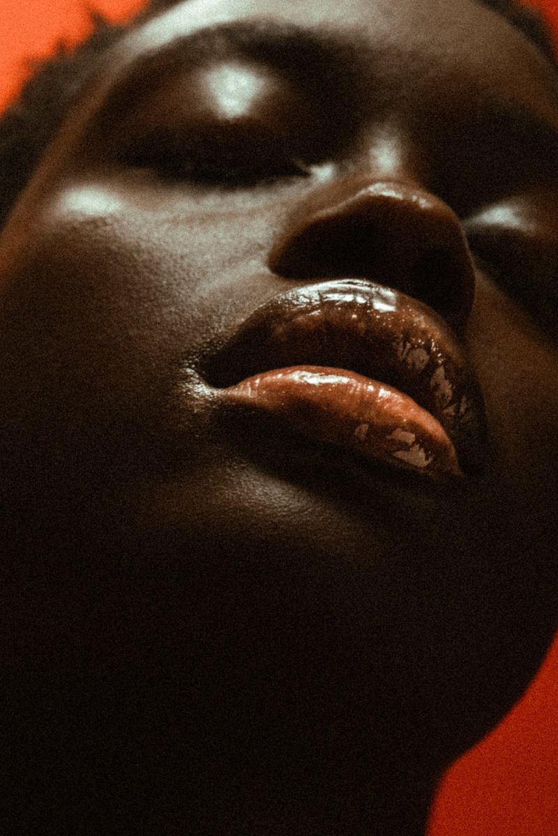 Close-up studio portrait of a dark-skinned woman with closed eyes, glossy lips, and highlighted cheekbones against an orange backdrop.