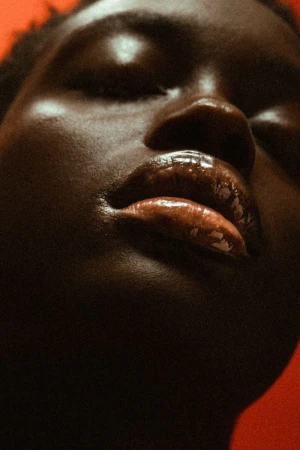 Close-up studio portrait of a dark-skinned woman with closed eyes, glossy lips, and highlighted cheekbones against an orange backdrop.