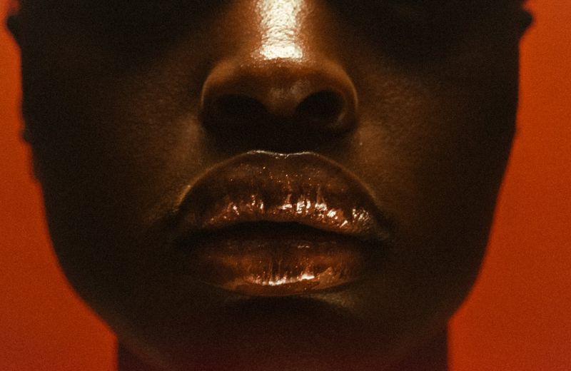 Close-up of an adult female's face, emphasizing glossy lips and dark skin tones, set against a solid orange backdrop. Studio lighting.