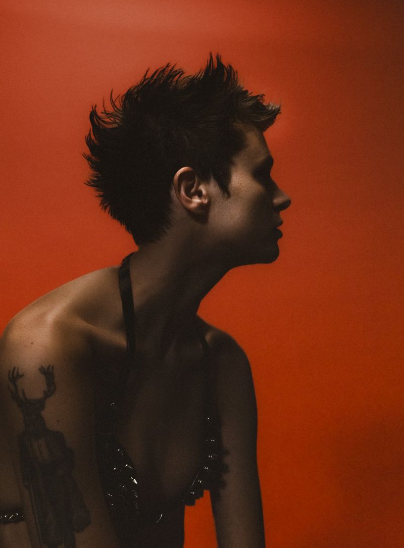 Close-up profile of a shirtless adult male with spiky dark hair against a plain orange background wearing a studded black top.