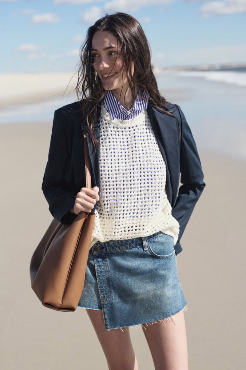 Brunette woman on beach in dark blazer, white sweater, and denim skirt carrying large tan tote bag, smiling with ocean in background.