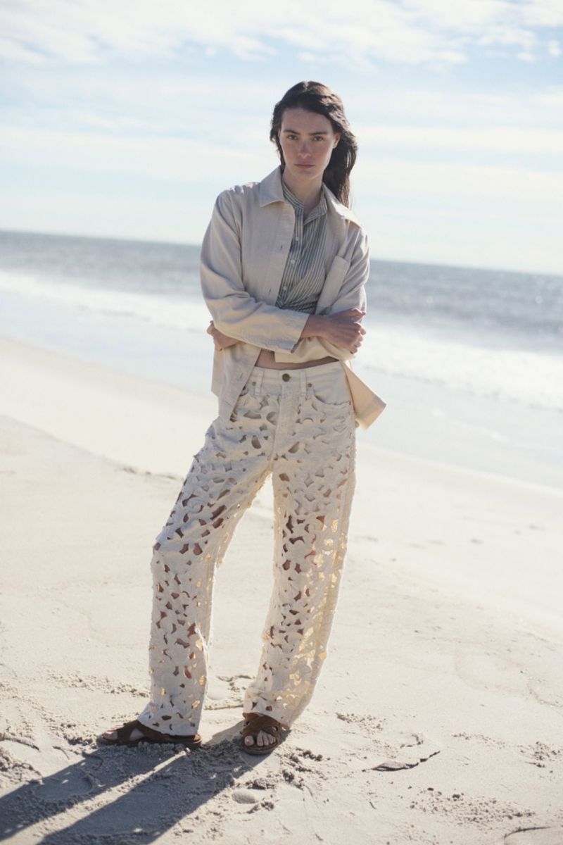 Woman poses on beach in fashion shot wearing cream-colored shirt and cut-out jeans against background of sand, ocean, and blue sky.