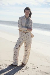 Model standing on a sandy beach wearing a cream jacket and patterned trousers against the ocean