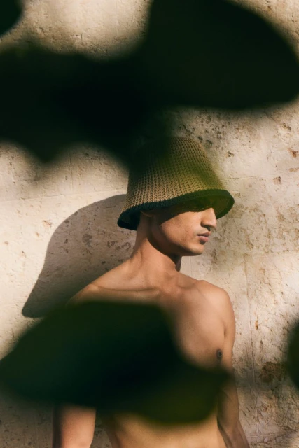 Adult male poses shirtless in a woven bucket hat with shadows, against a textured wall in sunlight, creating a natural fashion portrait.