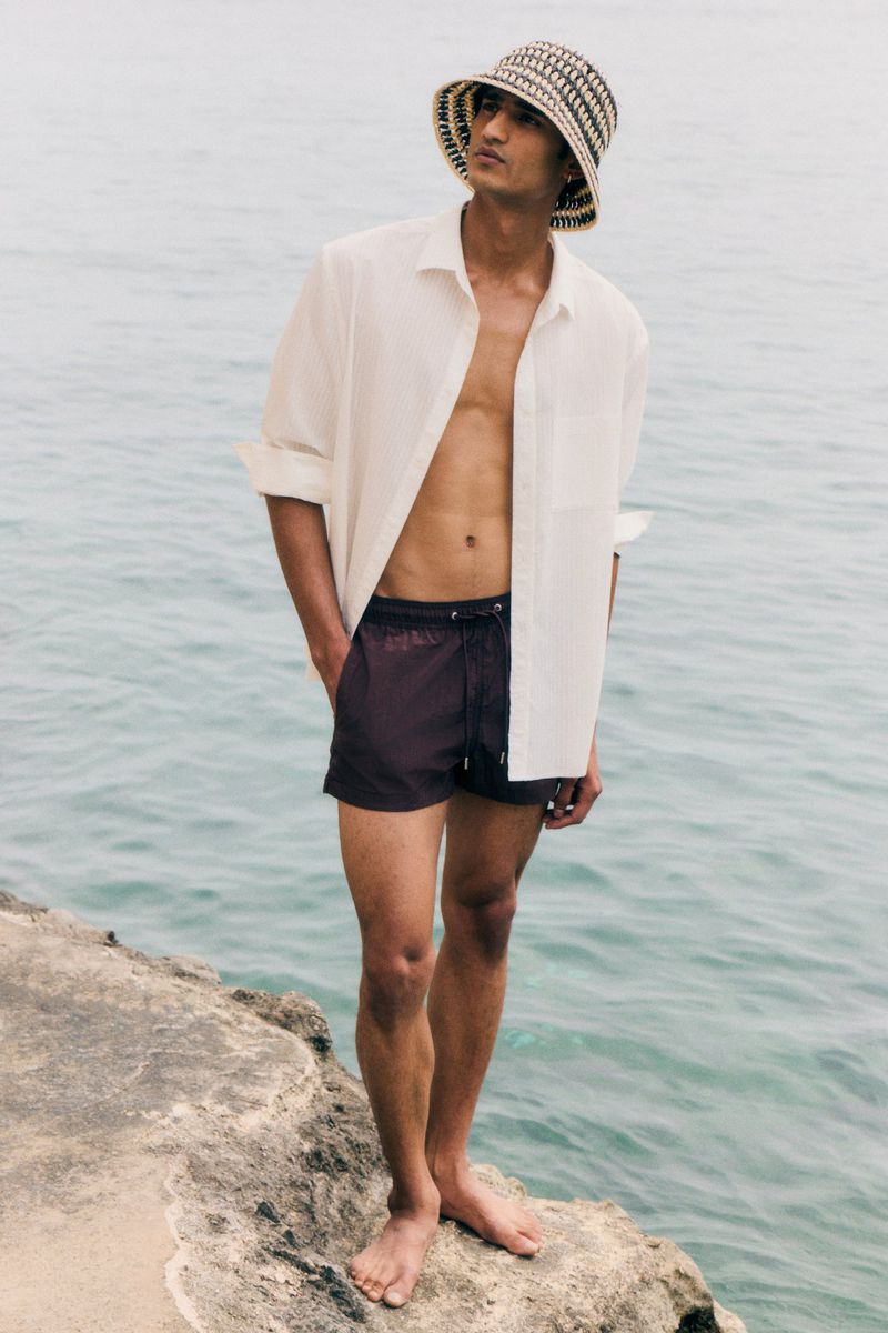 An adult male stands on a rocky outcrop wearing swim shorts, an unbuttoned shirt and bucket hat. The ocean stretches out behind him.