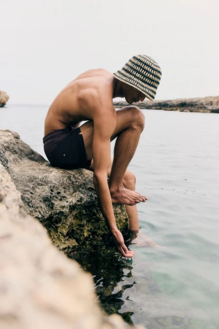 Young man sits on a rock at the beach, dipping his hand in the ocean water. He wears a striped bucket hat and dark swim trunks.