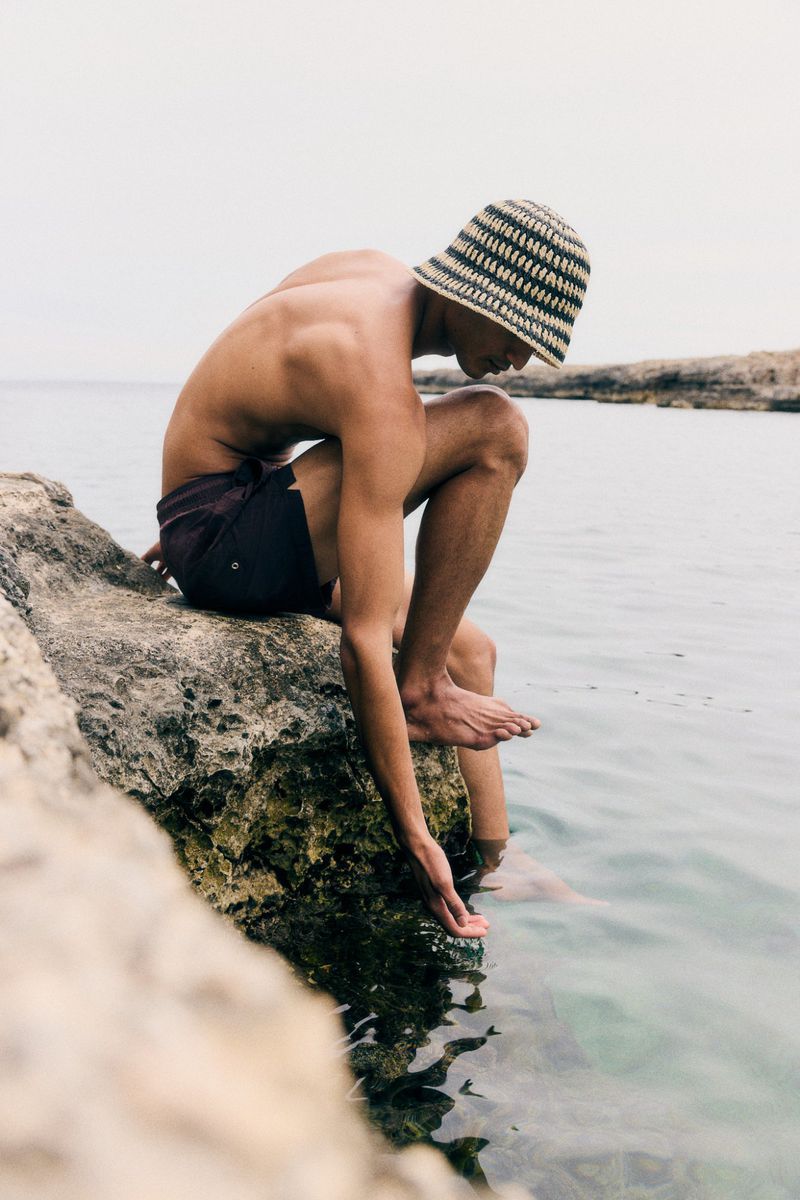 Man in swim trunks and striped hat sits on rocks at the water's edge, with hand touching clear water in an outdoor setting.
