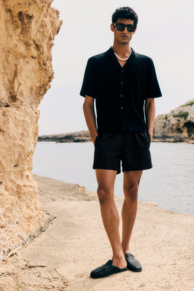 A stylish man poses on a stone surface near the sea, wearing a black button-down shirt, dark shorts, sunglasses, and slide shoes.