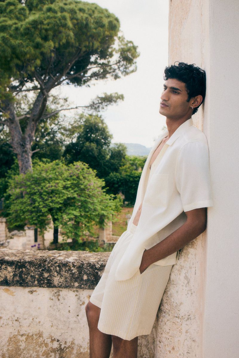 An adult male model poses outdoors near architecture with lush trees, wearing an open-collar shirt and shorts in a fashion editorial.