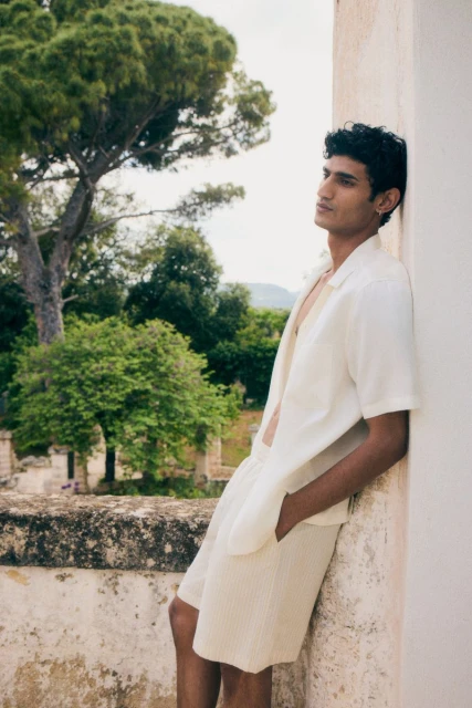 An adult male model poses outdoors near architecture with lush trees, wearing an open-collar shirt and shorts in a fashion editorial.
