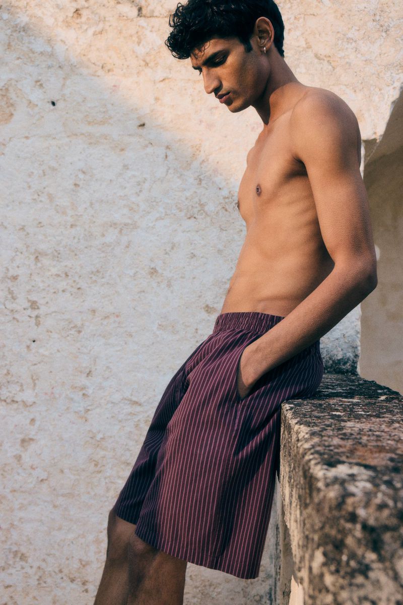 A shirtless man in striped shorts leans against a textured stone wall in outdoor setting with natural sunlight.