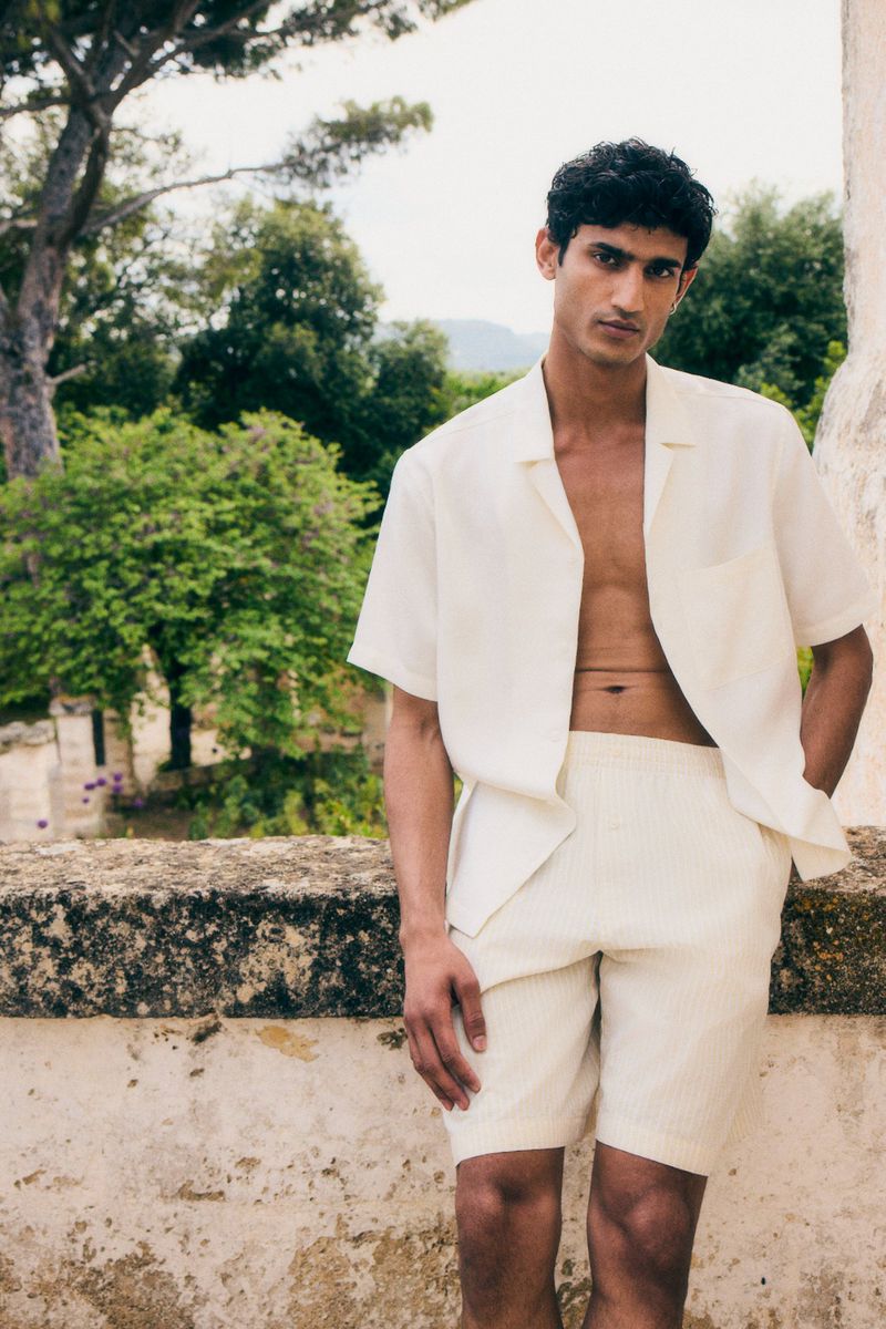 A man in white linen shorts and a matching short-sleeve shirt poses outdoors against a stone wall and a background of green trees.