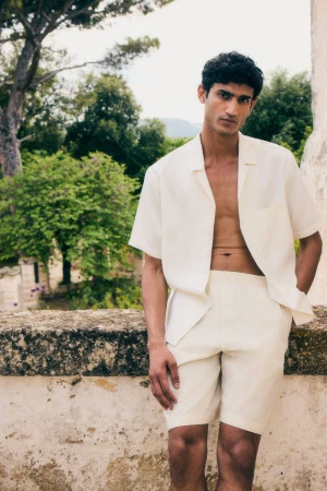 A man in white linen shorts and a matching short-sleeve shirt poses outdoors against a stone wall and a background of green trees.
