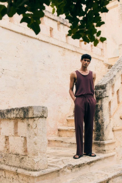 Man in casual maroon jumpsuit standing on light stone steps outdoors under tree branches, posing with hands in pockets.