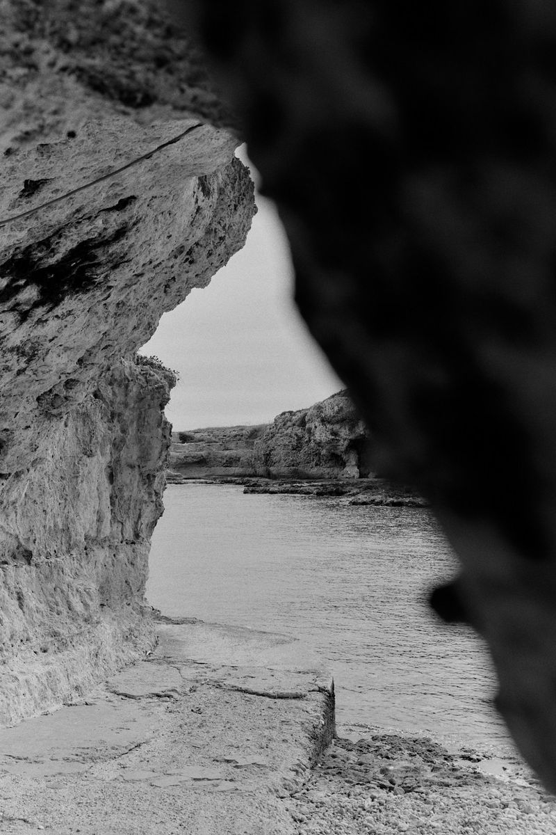 Black and white landscape of a rocky coast with cliffs and water, framed by rock formations in the foreground.