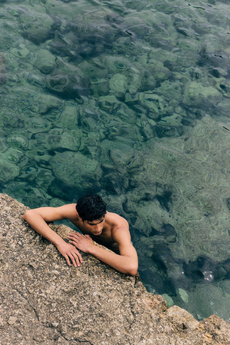 Adult male leans on rock in clear ocean water. His upper body is visible above the water with the rocky shore alongside.
