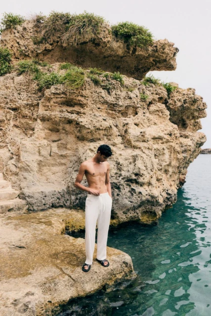 A dark-haired adult man stands on a rocky outcrop at the water's edge, wearing white trousers and sandals, with a rocky cliff behind.